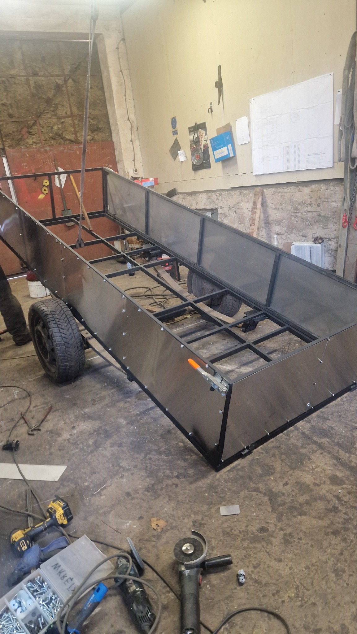 Metalowa przyczepa w trakcie budowy w warsztacie, widoczne spawane elementy ramy, poszycie, koła oraz narzędzia pracy.