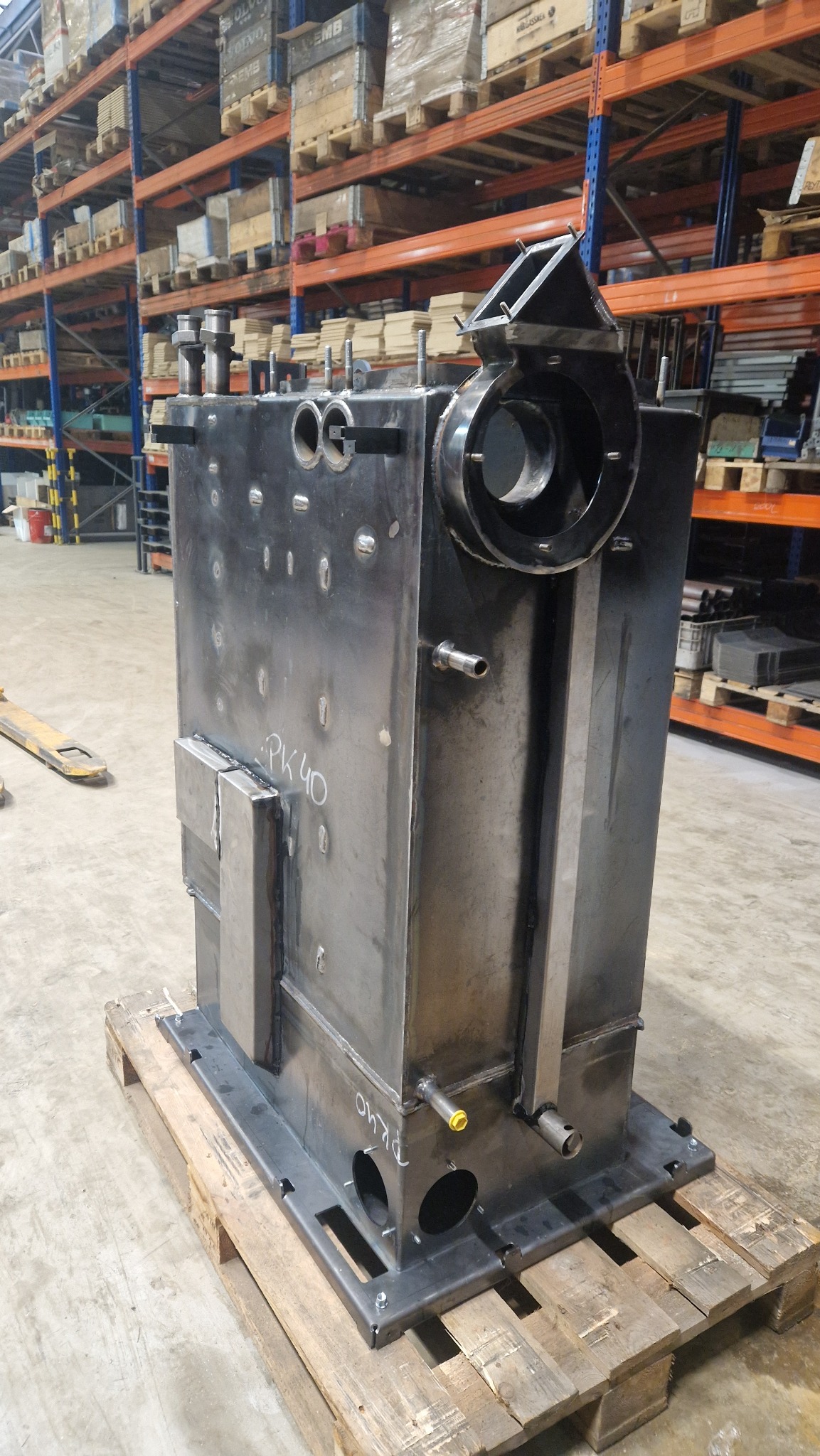 Surowy, metalowy zbiornik z widocznymi spawami i oznaczeniem PK40, umieszczony na drewnianej palecie w magazynie z regałami.