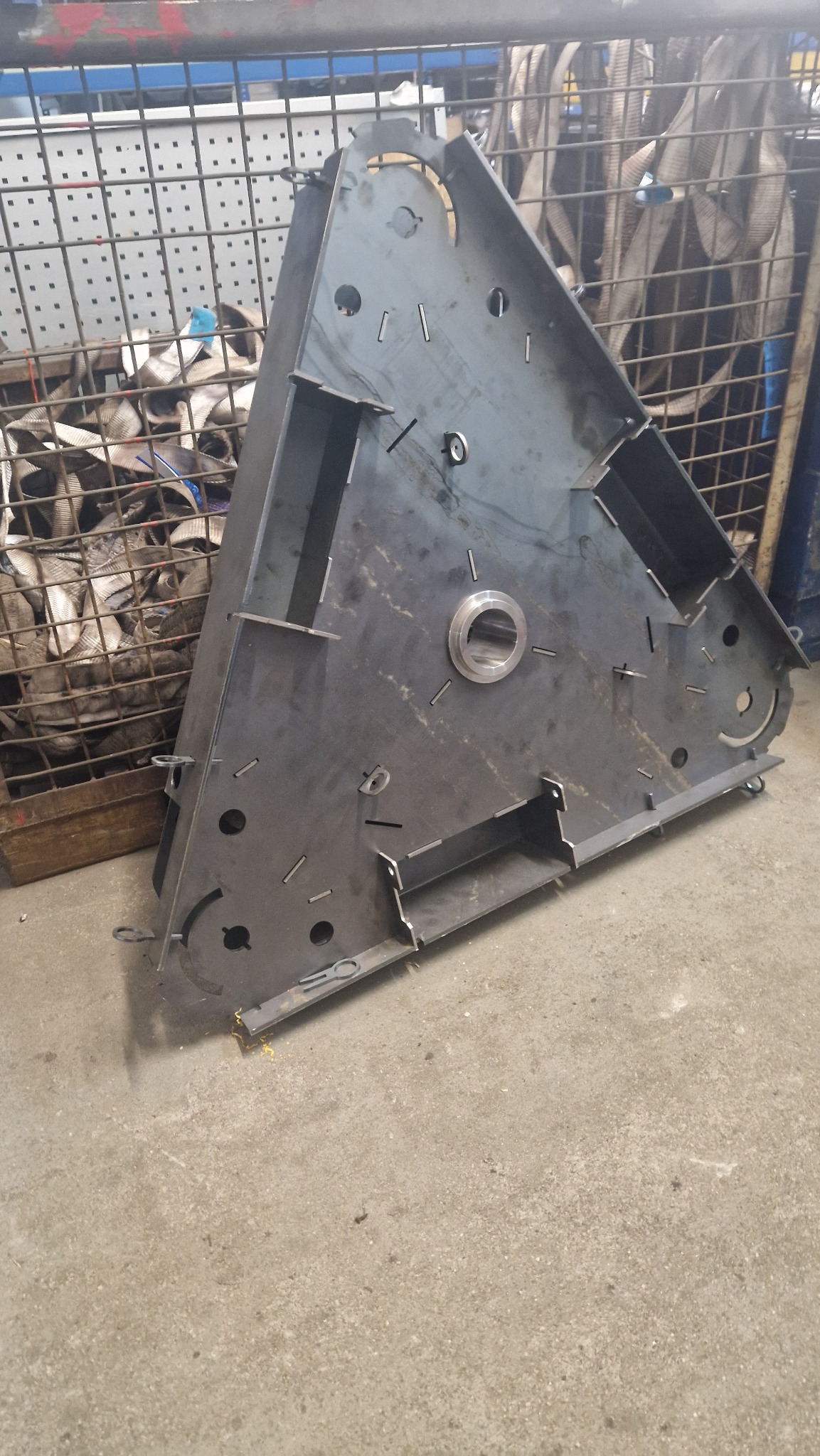 Trójkątna, metalowa konstrukcja z wyciętymi otworami i wspawanymi elementami, leżąca na betonowej podłodze w warsztacie, z widocznym koszem z pasami transportowymi w tle.