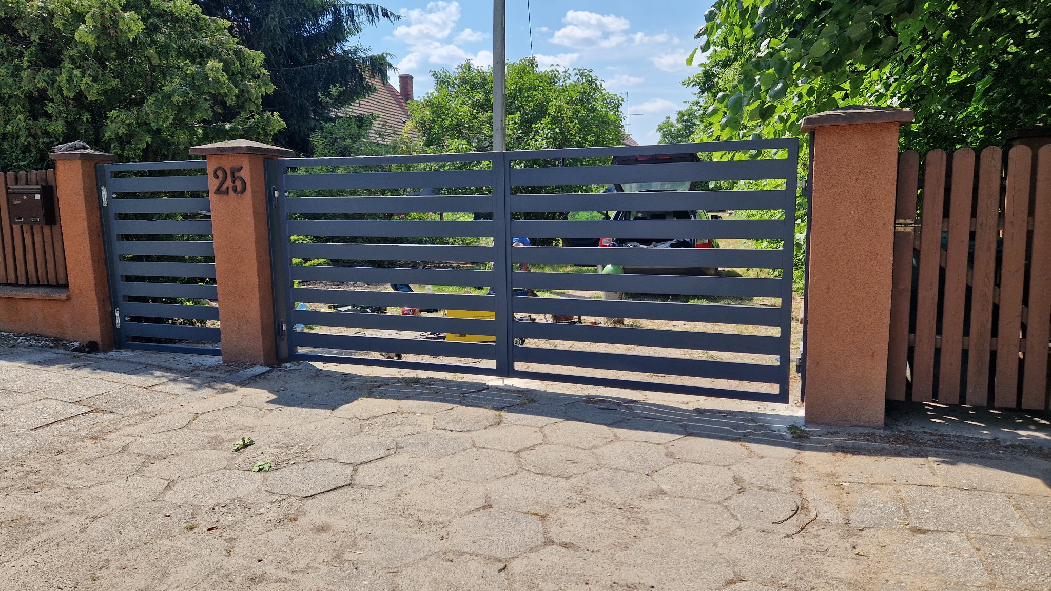 Nowoczesna, pozioma brama metalowa w kolorze grafitowym, zamontowana między ceglanymi słupkami z numerem domu '25', widoczne narzędzia i auto w tle, sugerujące trwający montaż.