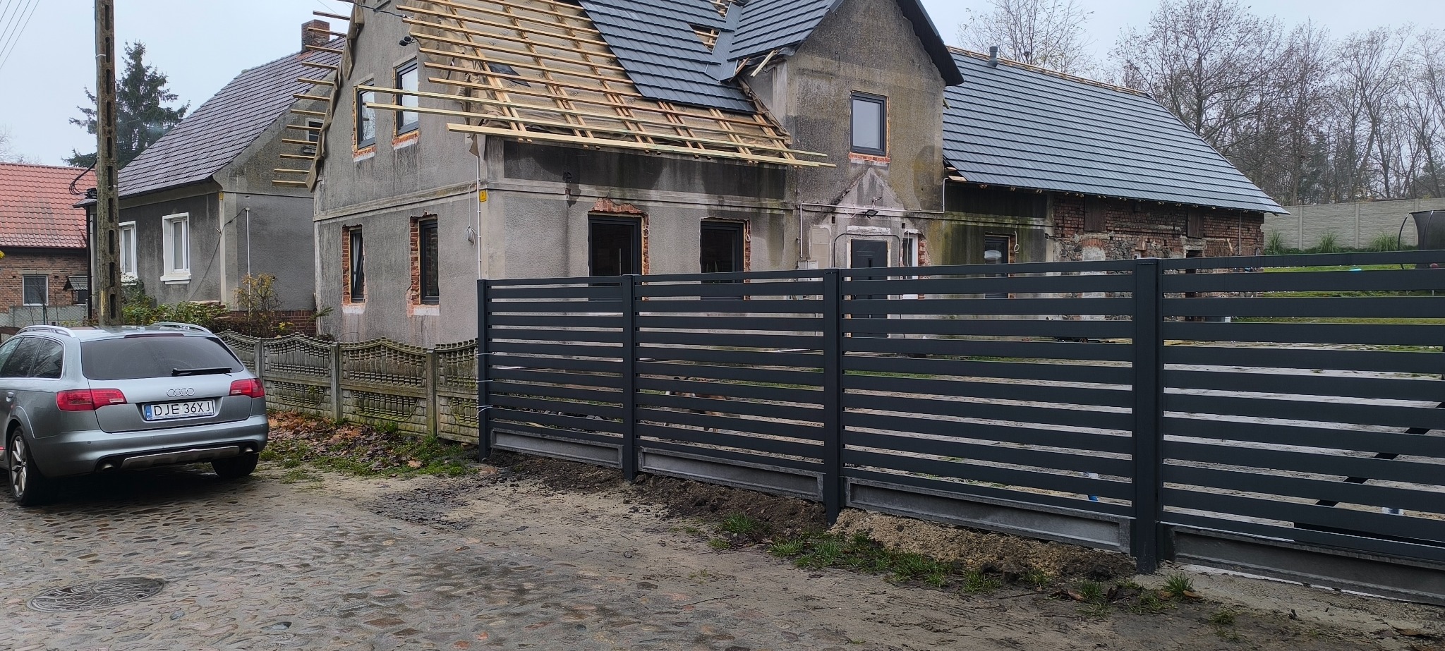 Nowoczesne, poziome ogrodzenie metalowe w kolorze grafitowym, na tle remontowanego domu z częściowo zdjętym dachem i szarym samochodem osobowym. Ujęcie z poziomu ulicy.