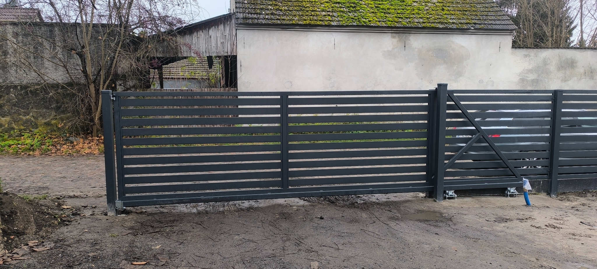 Nowoczesna, przesuwna brama wjazdowa z poziomych profili, szara, na tle budynku z mchem na dachu. Widoczne elementy mechanizmu przesuwnego.