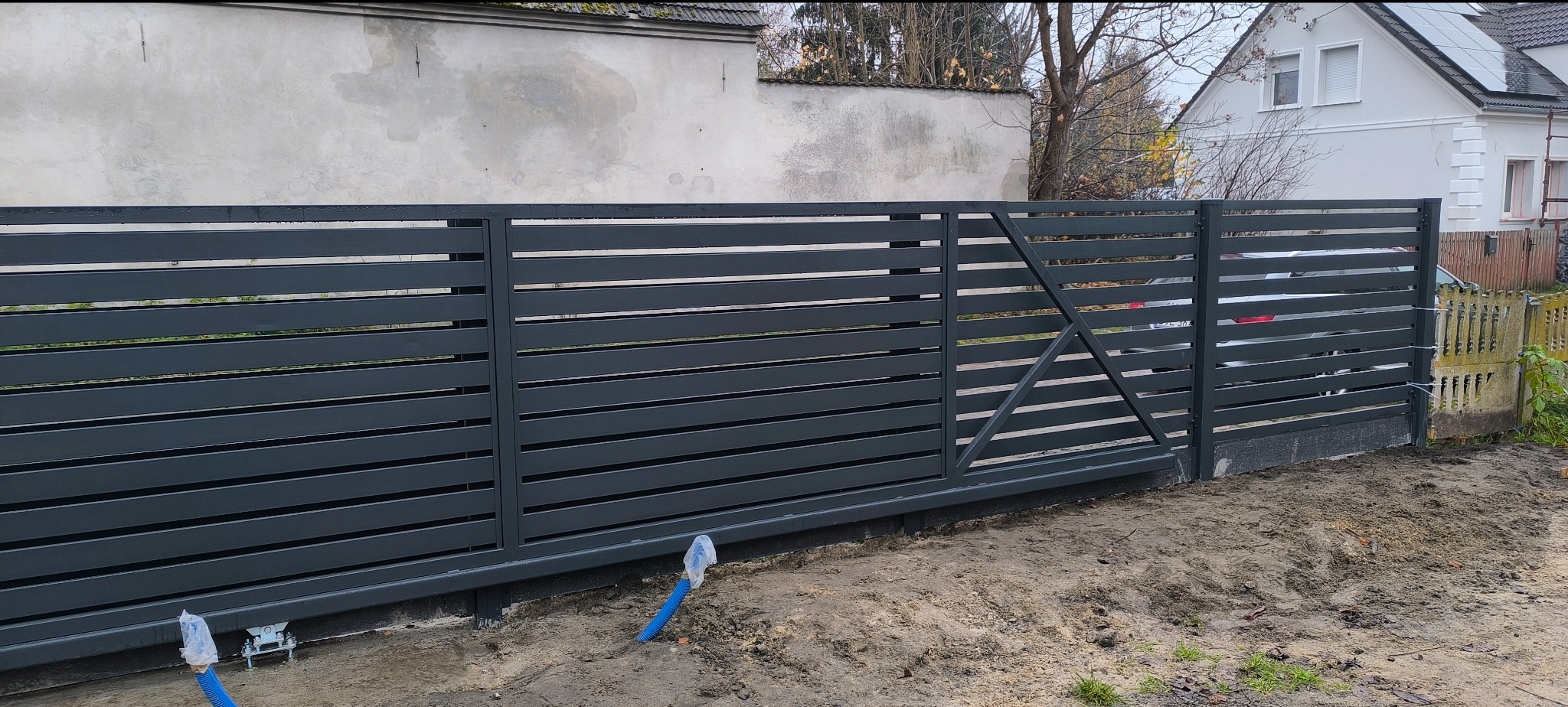 Nowoczesna, przesuwna brama wjazdowa z poziomych profili, pomalowana na grafitowo. Widoczne elementy montażowe i instalacja elektryczna w ziemi. Tło stanowi jasny budynek.