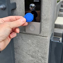 Novi-Tech Robert Nowicki - Ręka przykłada niebieski brelok RFID do nowoczesnego wideodomofonu HIKVISION z kamerą, zamontowanego na szarym, teksturowanym słupku przy wejściu.