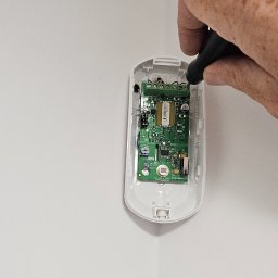 Novi-Tech Robert Nowicki - Instalator montuje urządzenie elektroniczne w narożniku pomieszczenia, widoczna otwarta obudowa z zieloną płytką drukowaną i dłoń z narzędziem do podłączania przewodów.