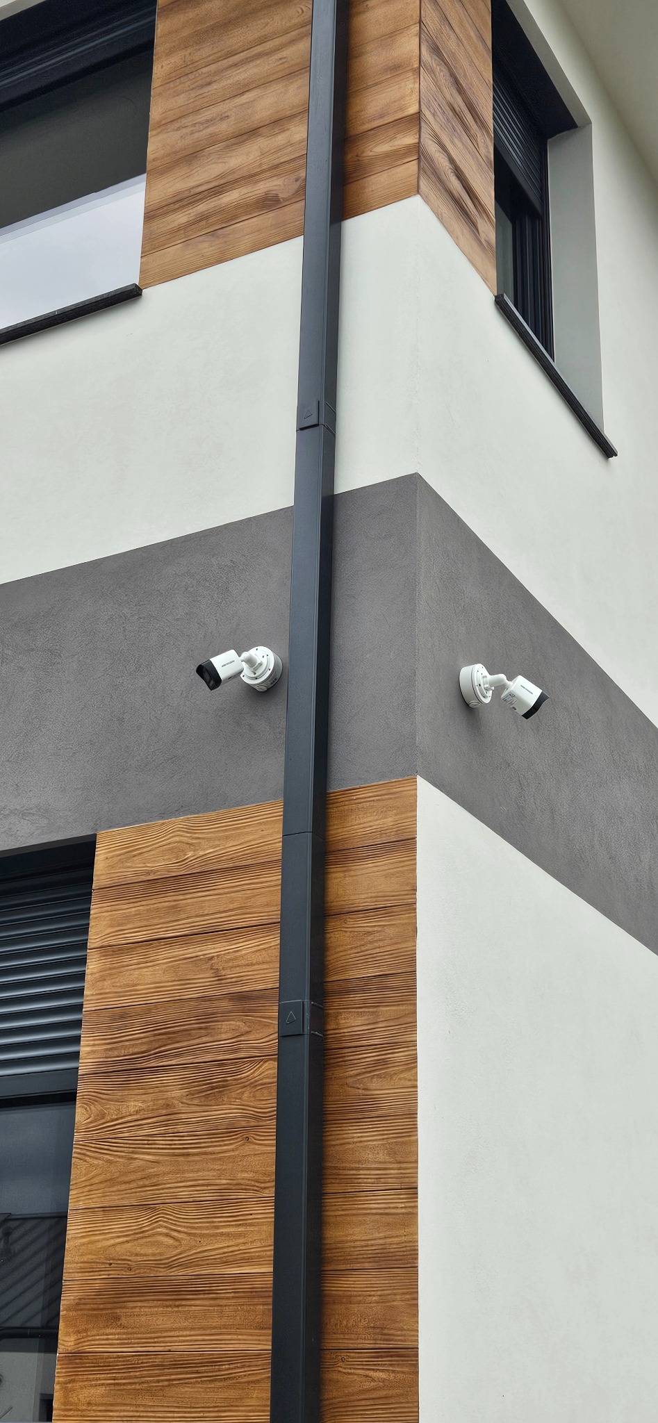 Dwie kamery monitoringu Hikvision zamontowane na elewacji budynku z drewnianą okładziną i szarą strefą. Widoczna rynna pionowa.