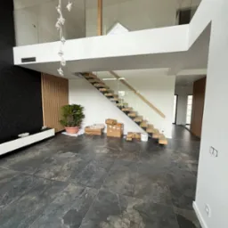 Nowoczesne wnętrze z przeszkloną balustradą na antresoli i drewnianymi schodami. Widok na otwartą przestrzeń z czarną ścianą i rośliną doniczkową.