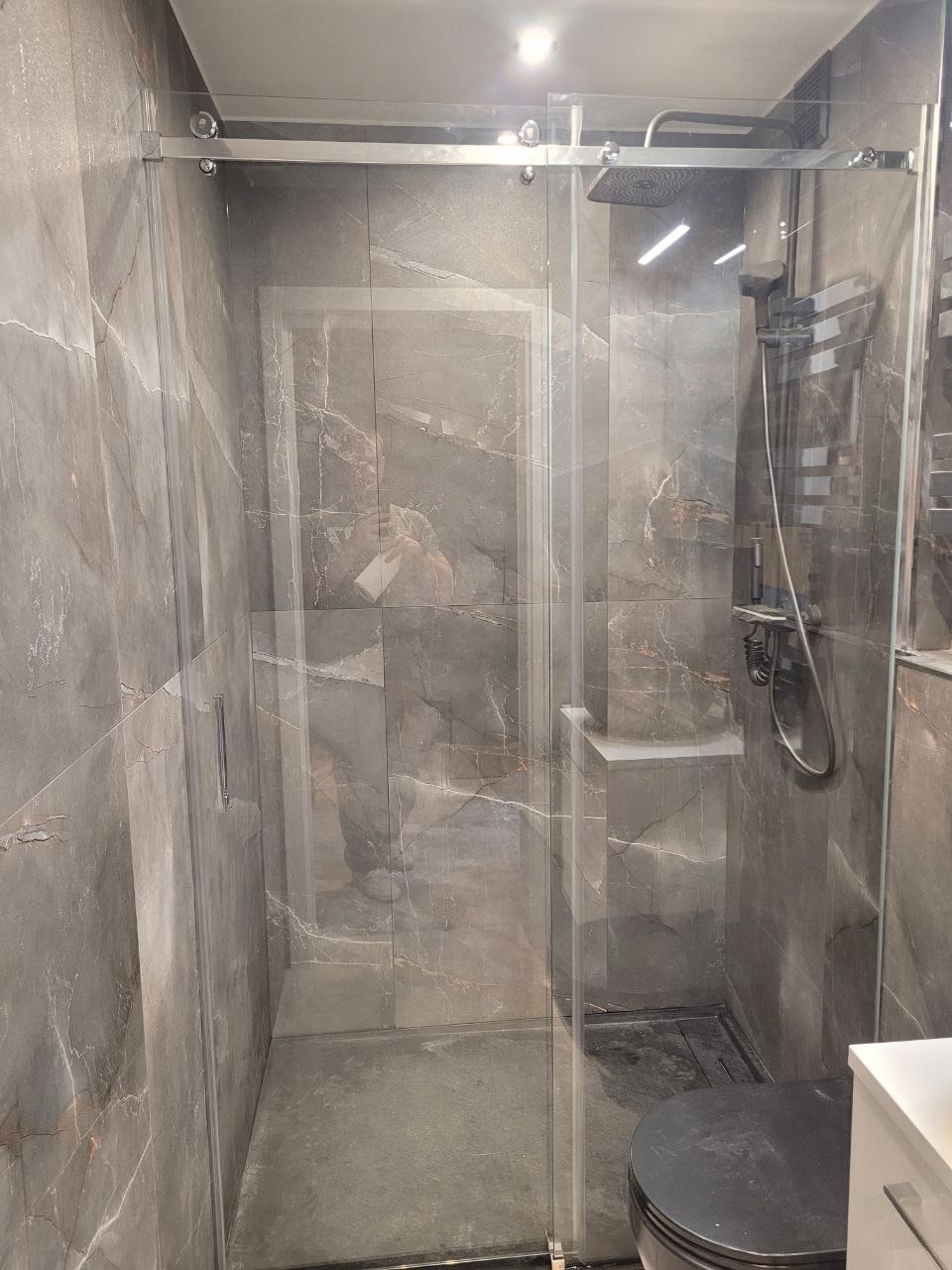 Kabina prysznicowa z przesuwnymi drzwiami szklanymi, wykończona płytkami imitującymi marmur w odcieniach szarości, widoczny zestaw natryskowy z deszczownicą i bateria.