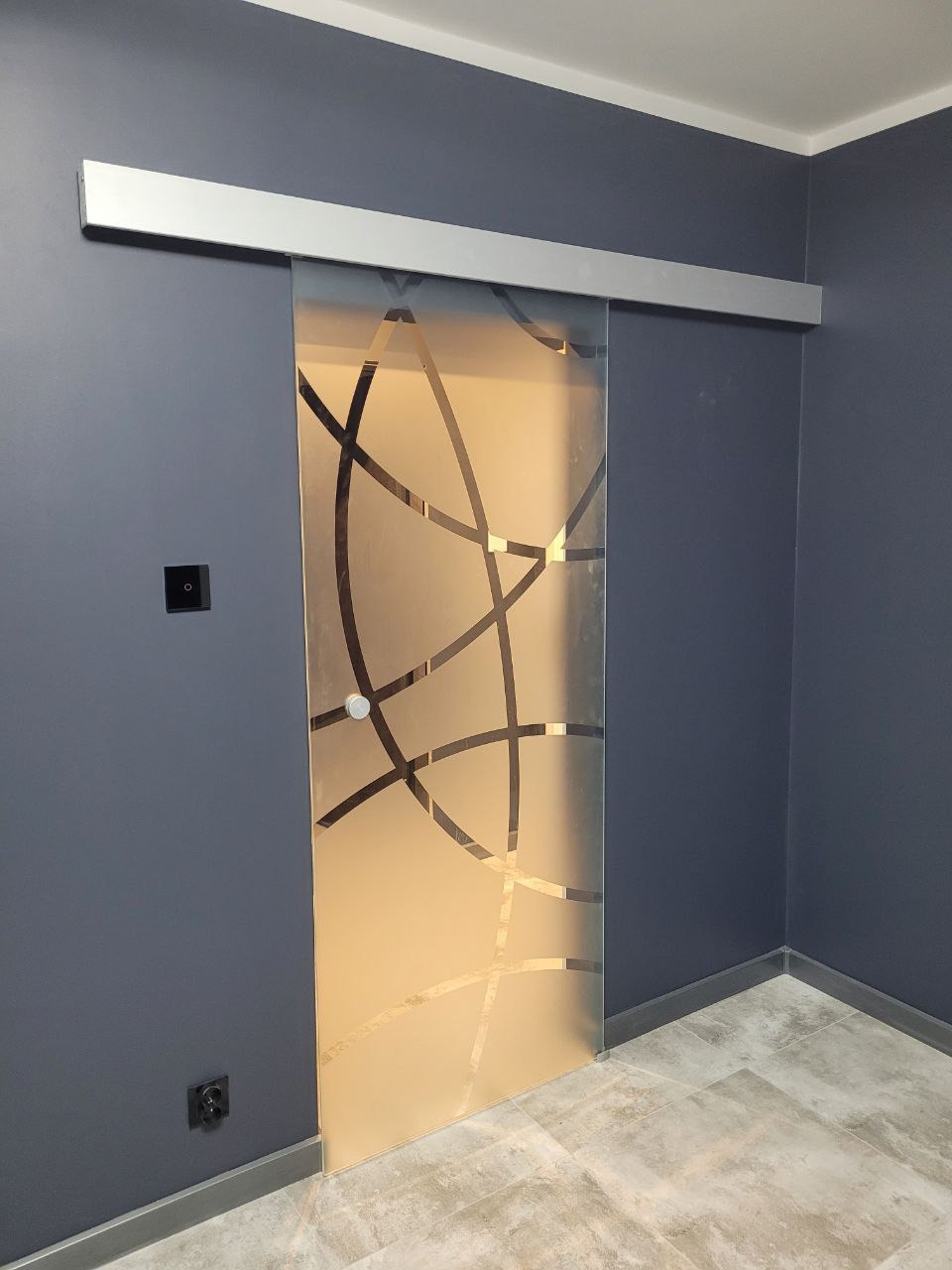 Szklane drzwi przesuwne z matowym wykończeniem i abstrakcyjnym wzorem geometrycznym, zamontowane na srebrnej szynie w pomieszczeniu z szarymi ścianami.