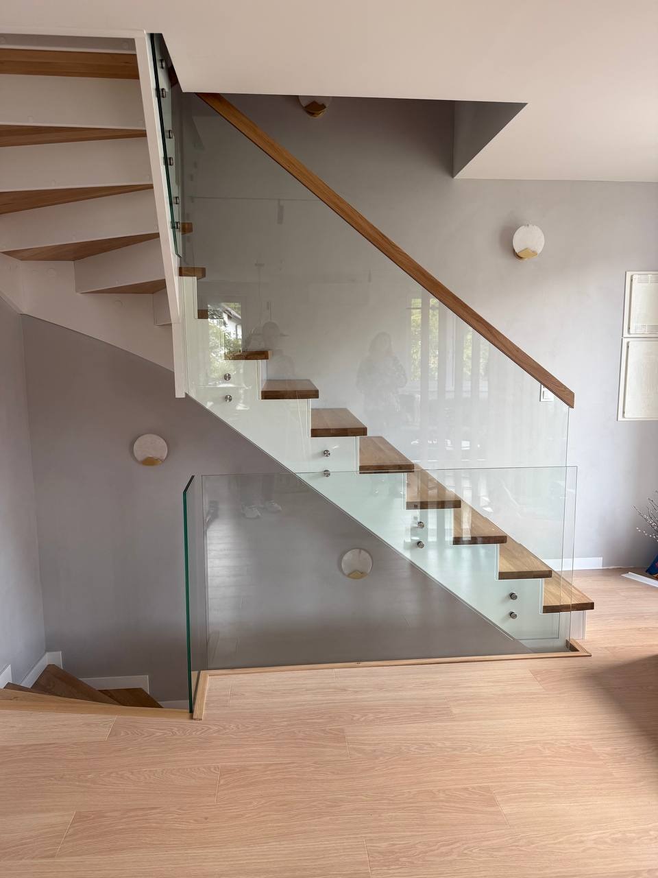Nowoczesna balustrada szklana ze schodami drewnianymi i minimalistycznymi lampami ściennymi w szarej tonacji, widok z dołu.