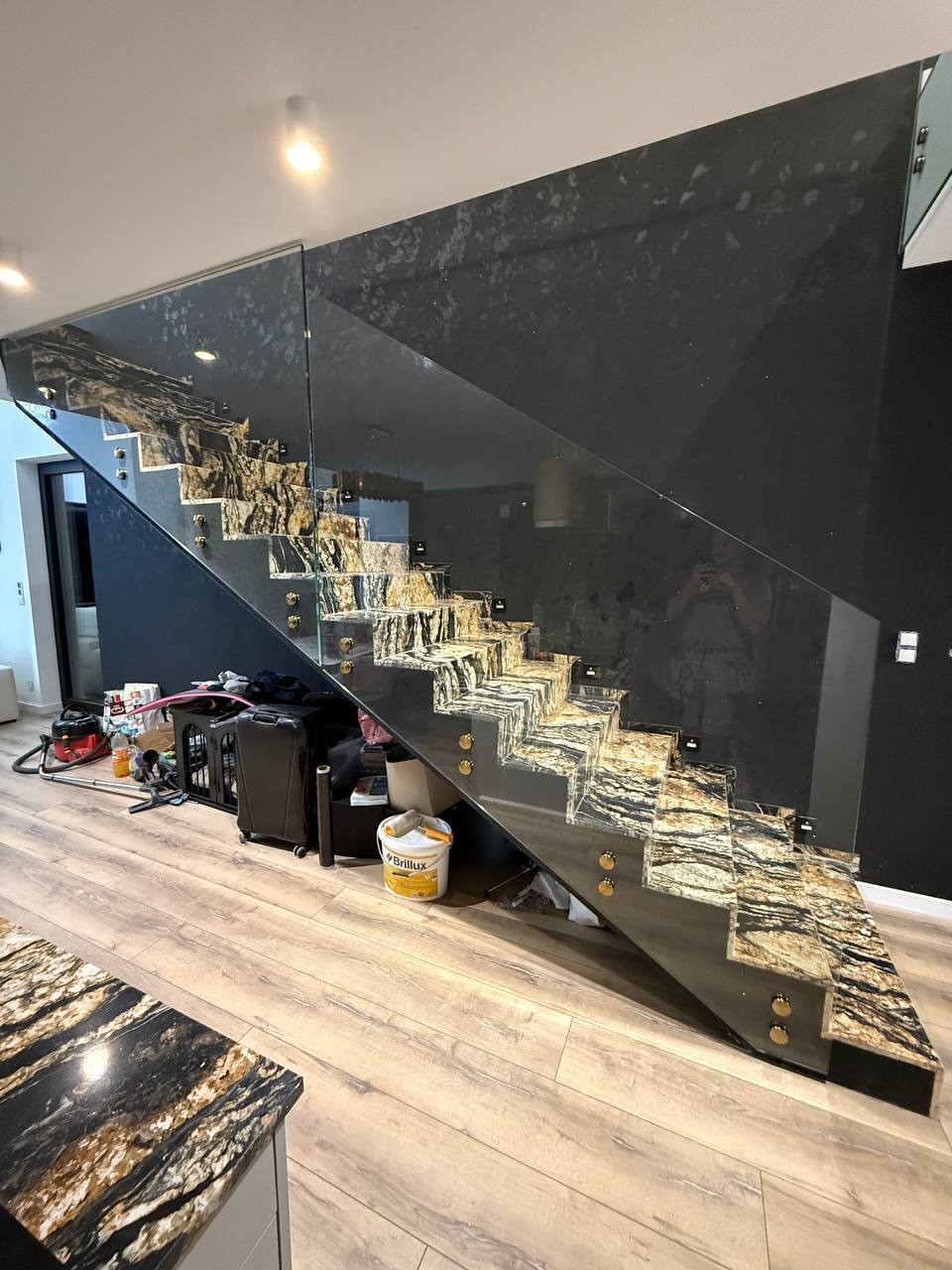 Nowoczesne schody z kamiennymi stopniami i szklaną balustradą, mocowaną na złotych elementach. W tle czarna ściana z dekoracyjną fakturą. Część blatu widoczna w lewym dolnym rogu.