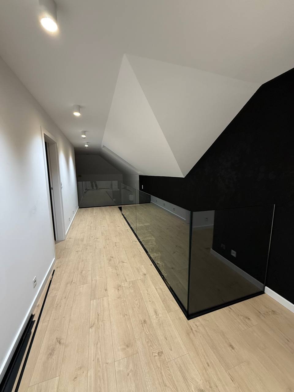 Nowoczesne, minimalistyczne wnętrze z szklaną balustradą wzdłuż korytarza. Jasne drewniane panele kontrastują z czarną ścianą i czarnymi elementami balustrady.