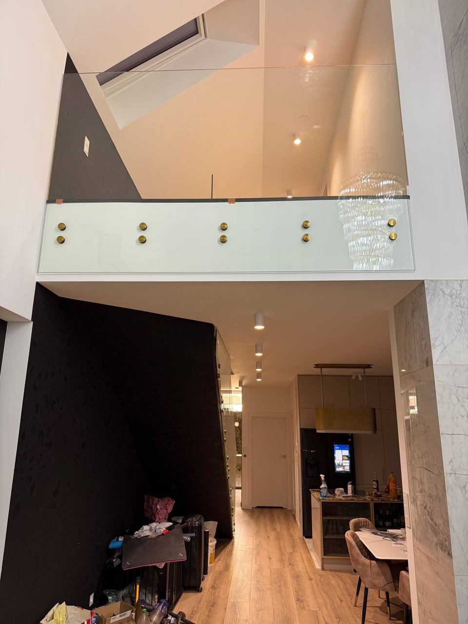 Nowoczesna balustrada szklana z mocowaniami w kolorze złotym oddzielająca przestrzeń salonu z widokiem na aneks kuchenny, oświetlona światłem dziennym.