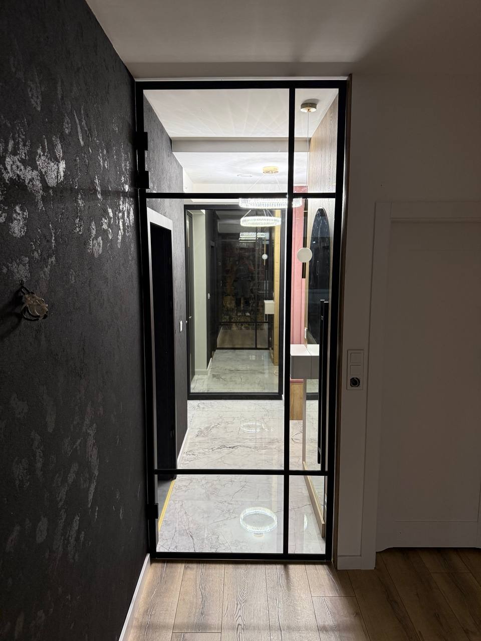 Nowoczesne drzwi szklane z czarną ramą, dzielące przestrzeń w domu. Widok na korytarz z marmurową podłogą i designerskim oświetleniem. Dekoracyjna ściana w tle.