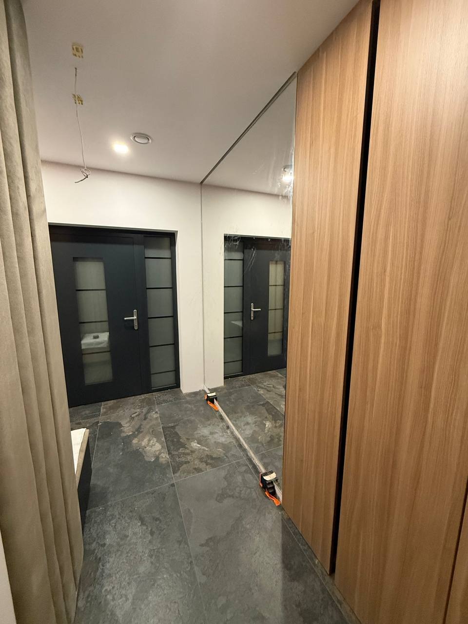 Nowoczesne, czarne drzwi szklane z podziałami w korytarzu z szarą podłogą. Widoczne narzędzia montażowe. Część ściany wykończona drewnem.