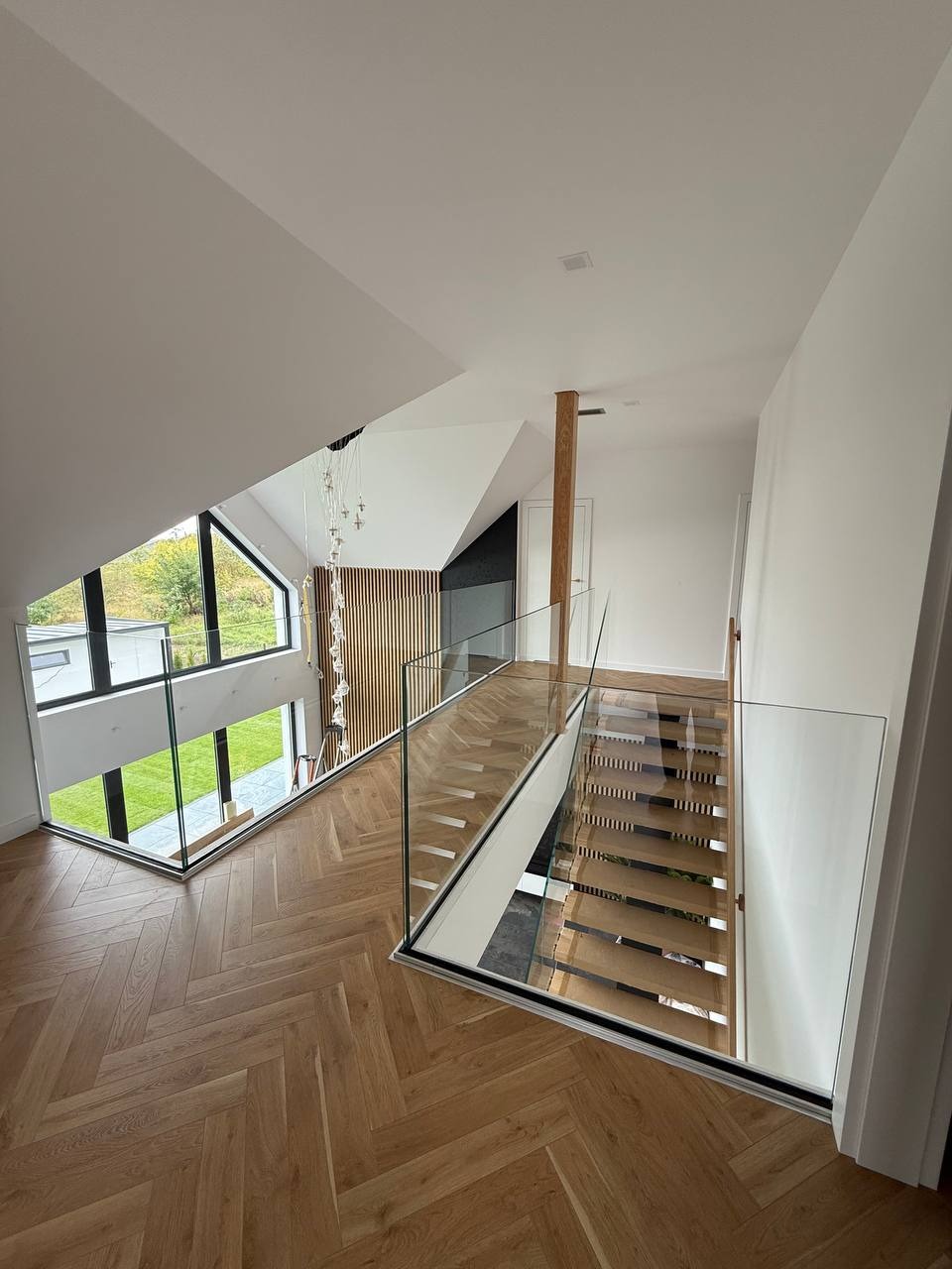 Szklana balustrada na antresoli z widokiem na schody i okno w nowoczesnym domu z parkietem w jodełkę. Minimalistyczny design z drewnianymi akcentami.