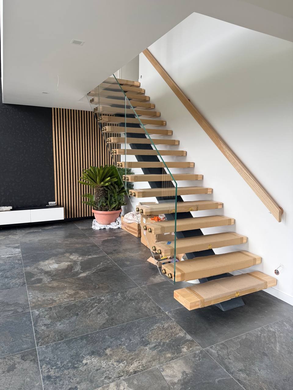 Nowoczesne schody z drewnianymi stopniami i szklaną balustradą, zabezpieczone kartonem, w jasnym wnętrzu z ciemną ścianą i rośliną doniczkową.