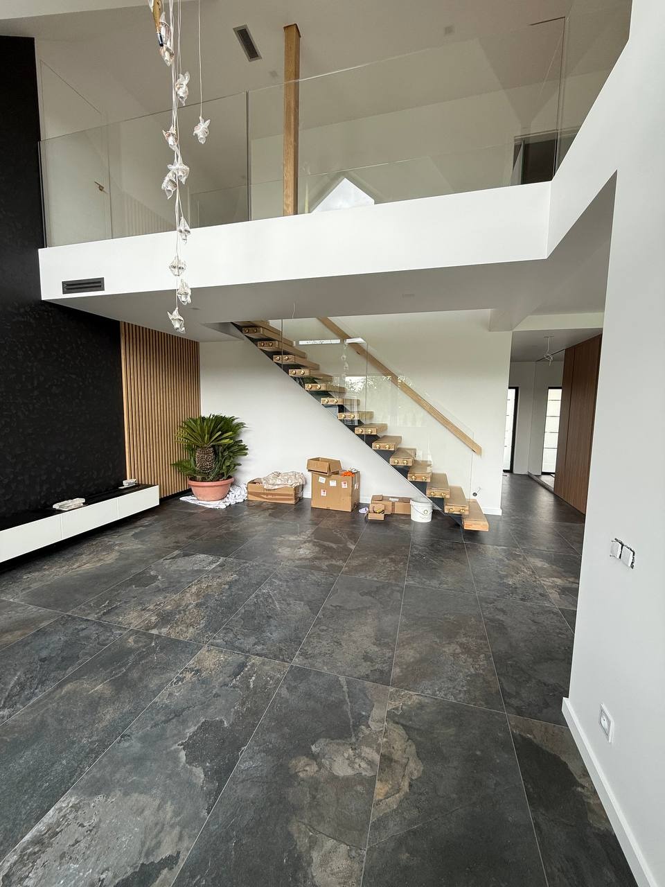 Nowoczesne wnętrze z przeszkloną balustradą na antresoli i drewnianymi schodami. Widok na otwartą przestrzeń z czarną ścianą i rośliną doniczkową.