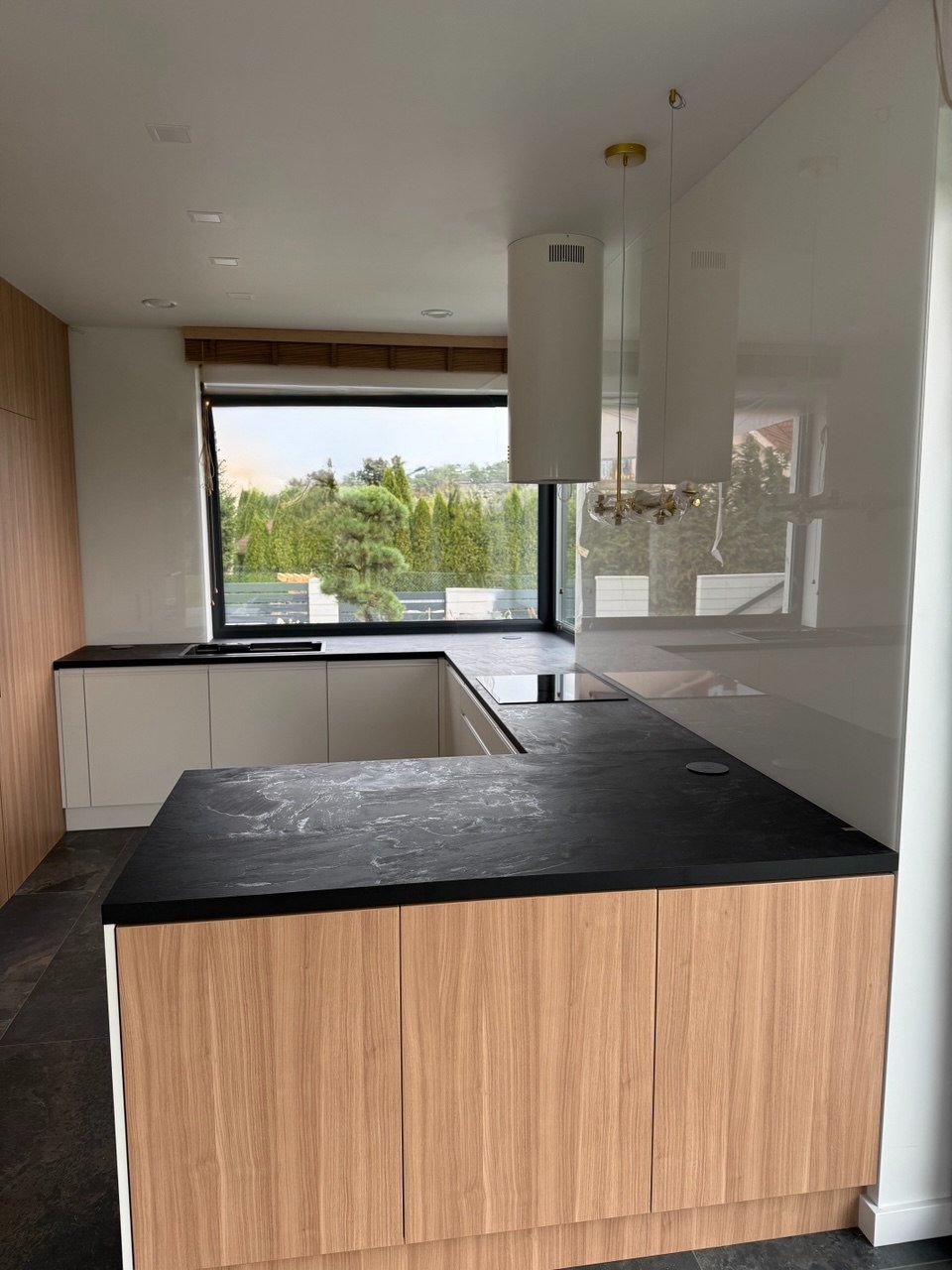 Nowoczesna kuchnia z dużym oknem i czarnym blatem. Minimalistyczny design, jasne szafki, widok na zieleń za oknem. Lampa wisząca z elementami złota.