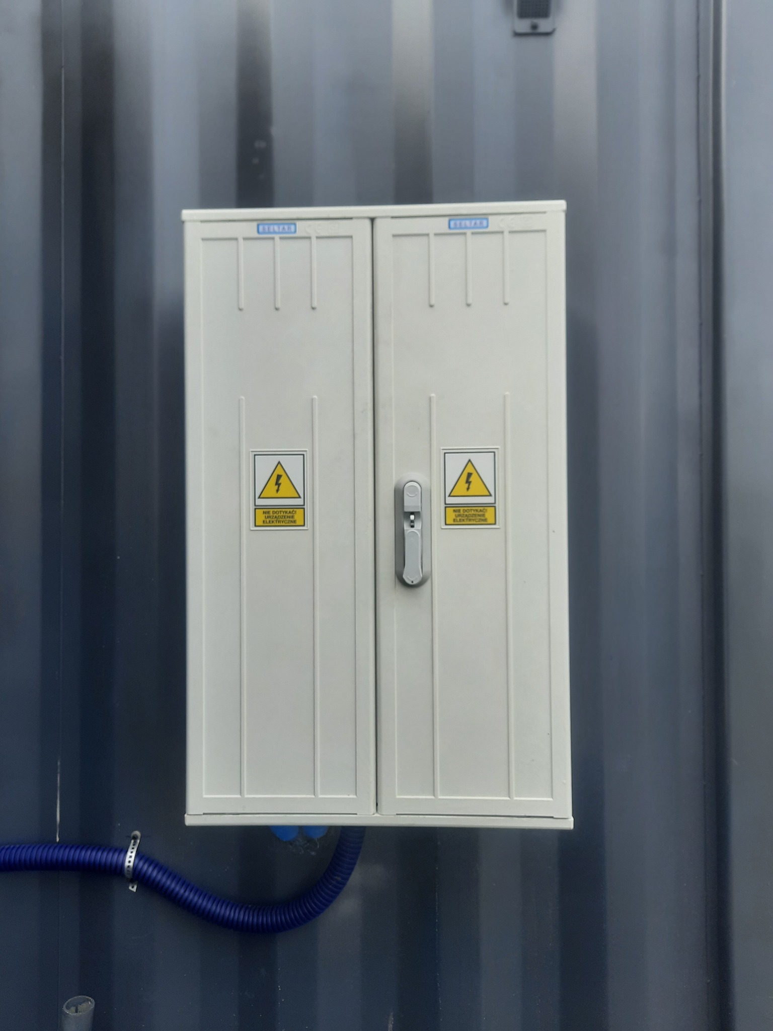 Biała skrzynka elektryczna z dwoma drzwiczkami, oznaczona żółtymi naklejkami ostrzegawczymi, zamontowana na ciemnoszarej ścianie kontenera, z widoczną niebieską rurą karbowaną doprowadzoną od dołu.