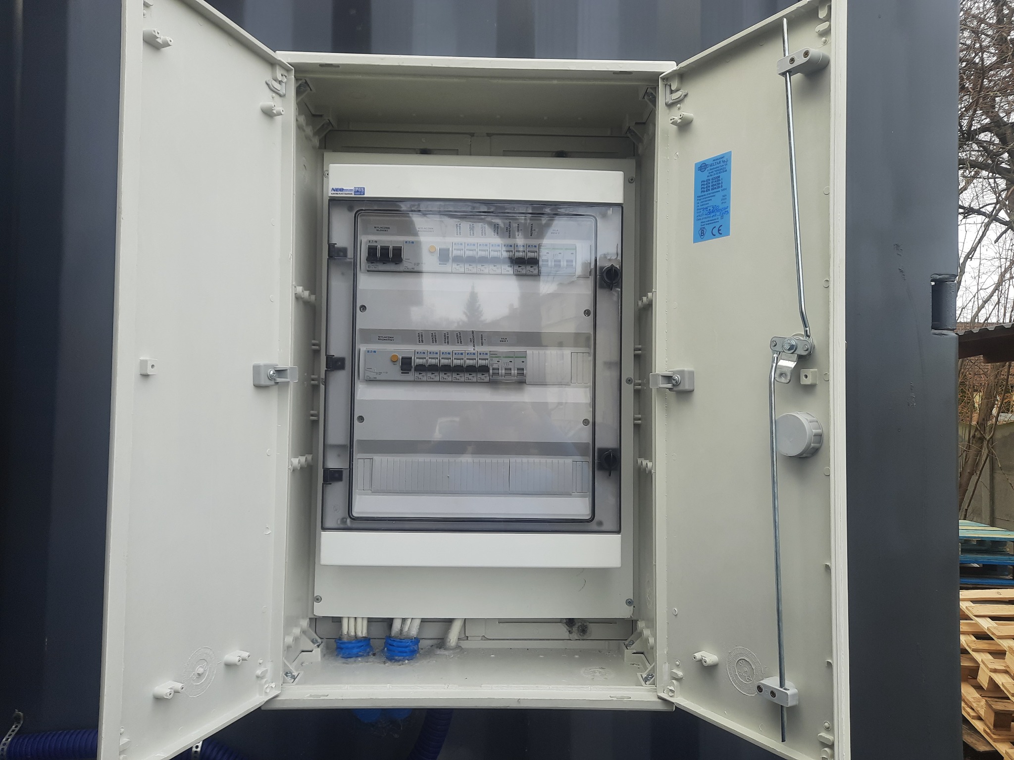 Otwarta skrzynka rozdzielcza na zewnątrz kontenera, widoczne zabezpieczenia, kable w niebieskich peszlach, tabliczka znamionowa na drzwiach.