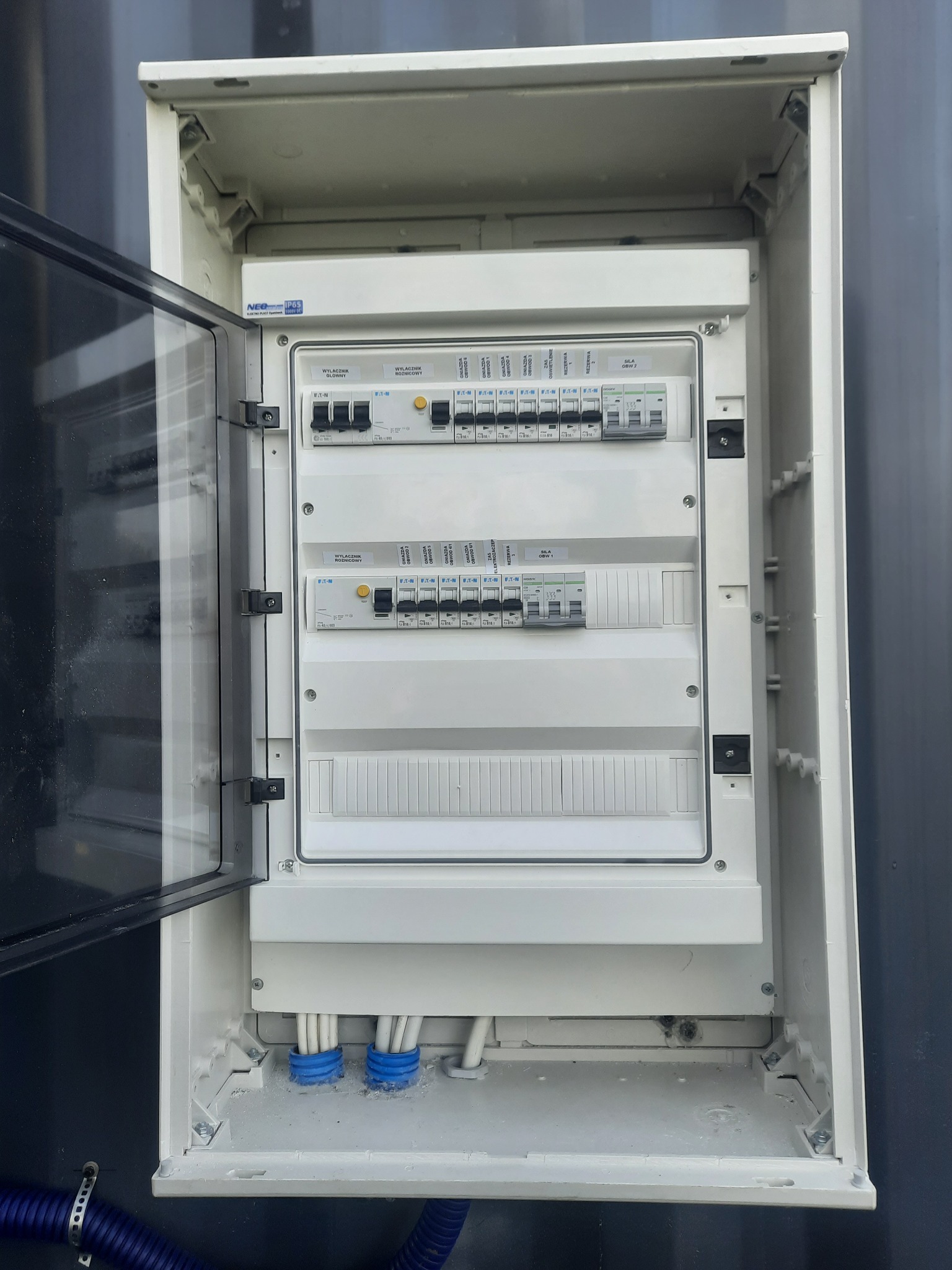 Otwarta, biała skrzynka rozdzielcza z widocznymi bezpiecznikami, wyłącznikami różnicowoprądowymi i wchodzącymi kablami w niebieskich peszlach, zamontowana na szarej ścianie.