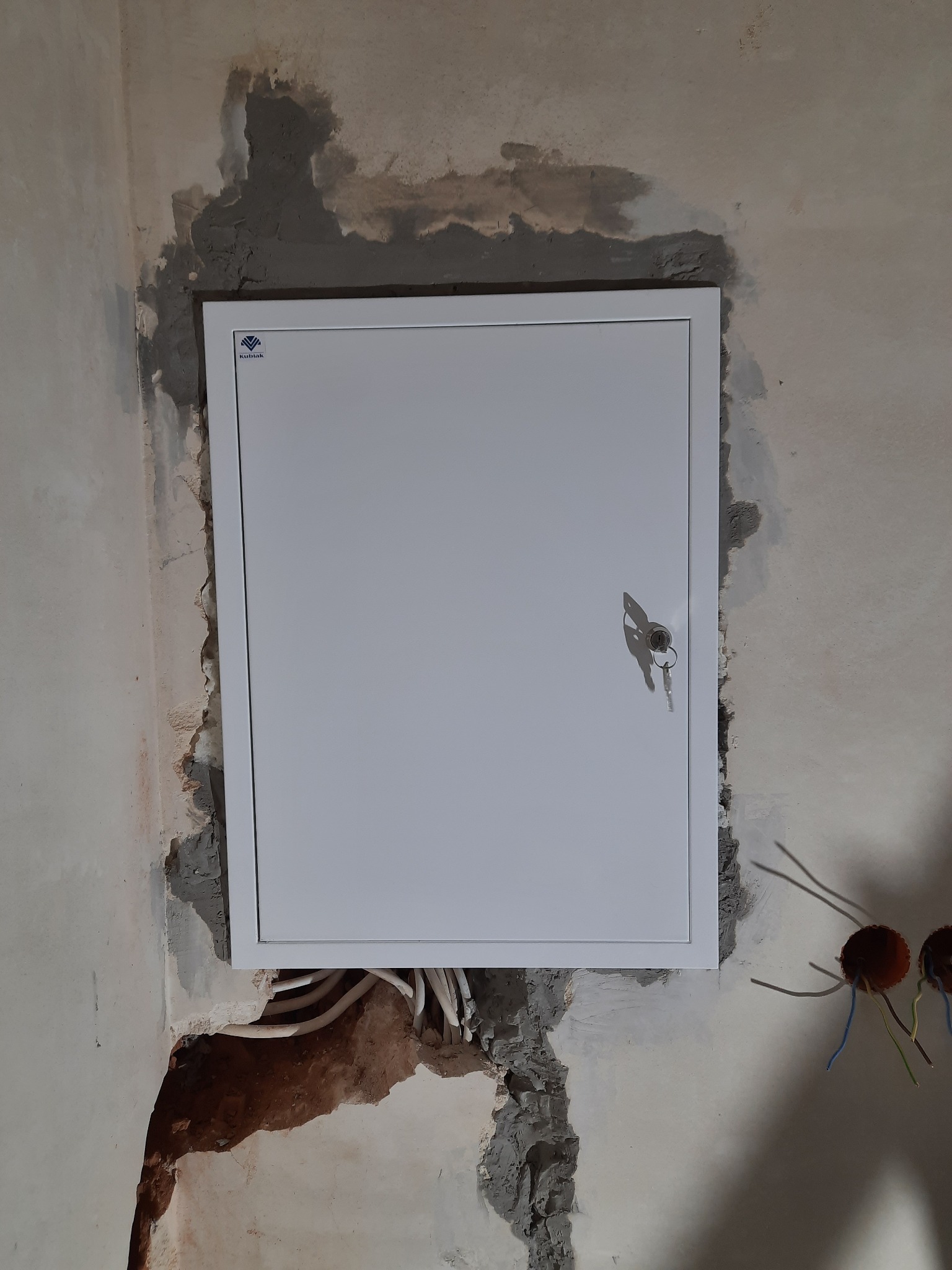 Biała szafka elektryczna z kluczykiem w zamku, zamontowana w surowej ścianie z widocznymi przewodami i śladami zaprawy.