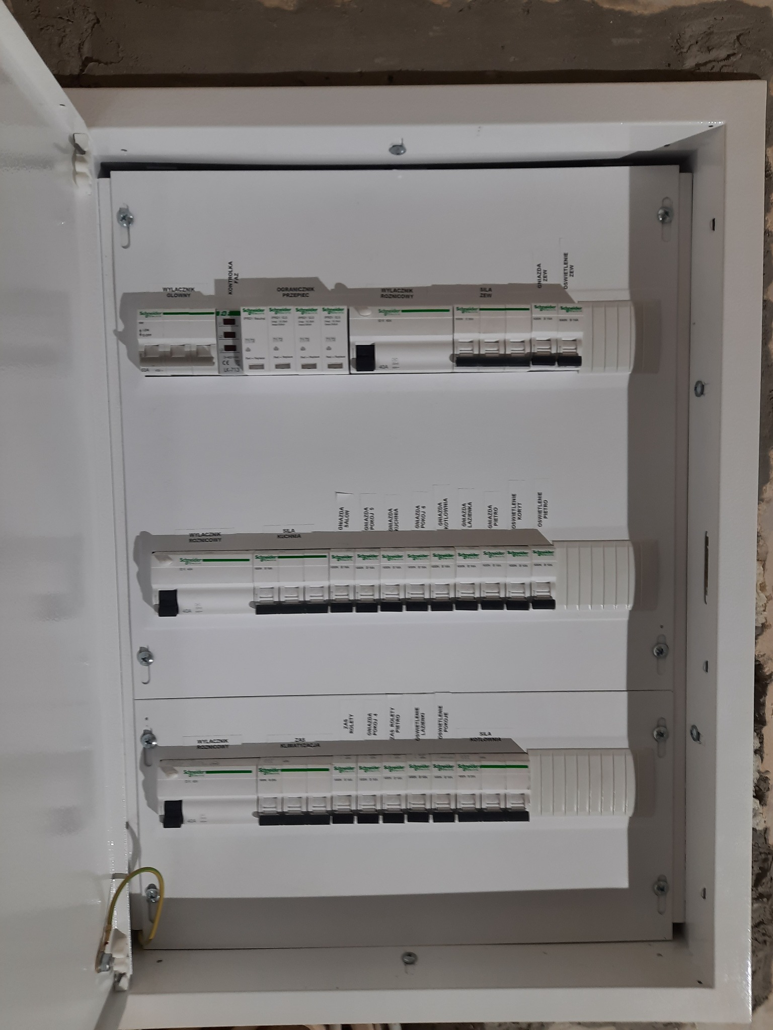 Otwarta skrzynka rozdzielcza z zainstalowanymi wyłącznikami różnicowoprądowymi i nadprądowymi marki Schneider, widoczne okablowanie ochronne, surowy mur w tle.