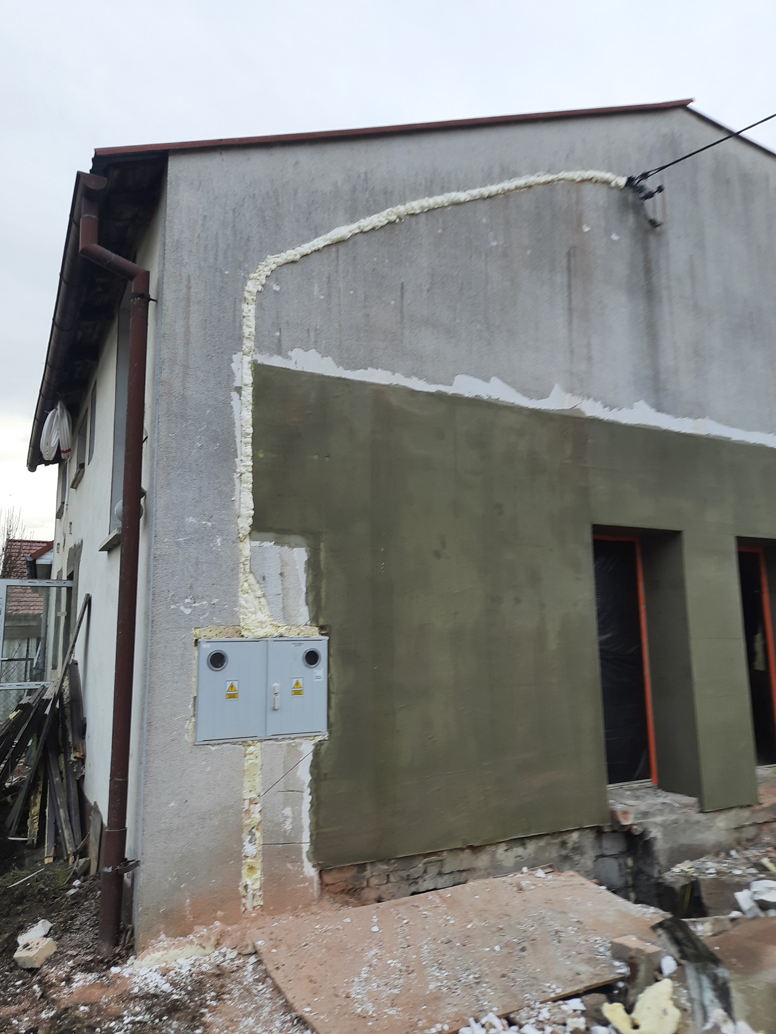 Ściana budynku w trakcie ocieplania z widoczną skrzynką elektryczną, pianką montażową i świeżo nałożonym tynkiem w kolorze oliwkowym.