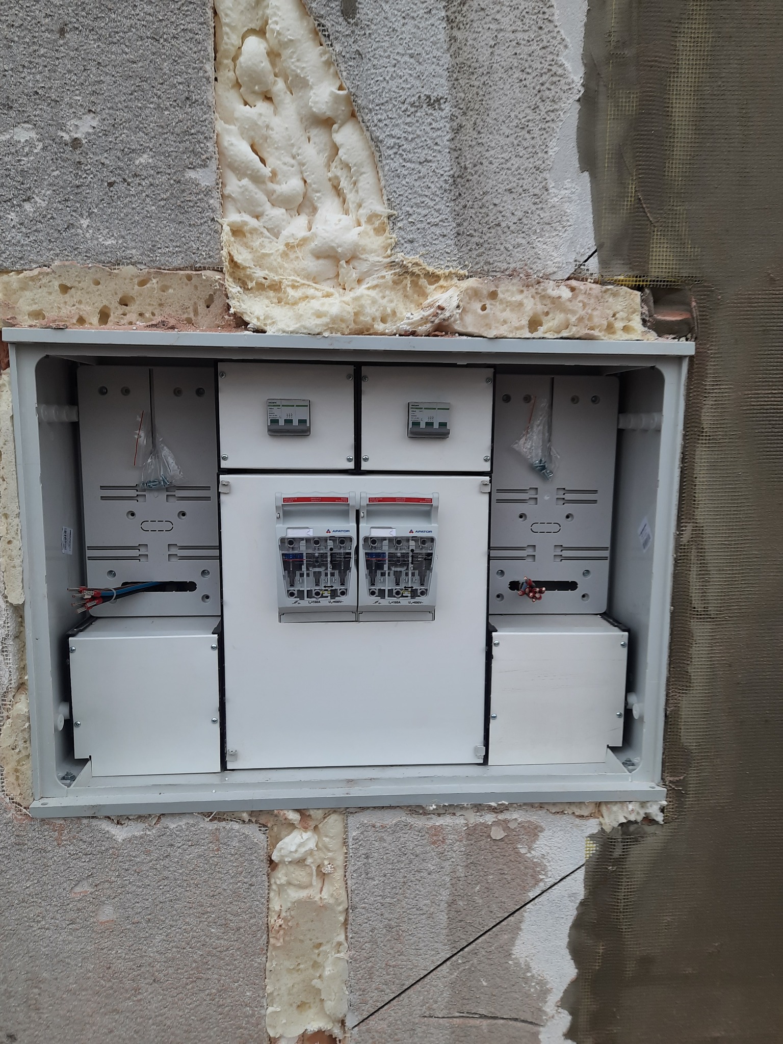 Skrzynka elektryczna z zabezpieczeniami w trakcie montażu w surowej ścianie z widoczną izolacją i siatką podtynkową.
