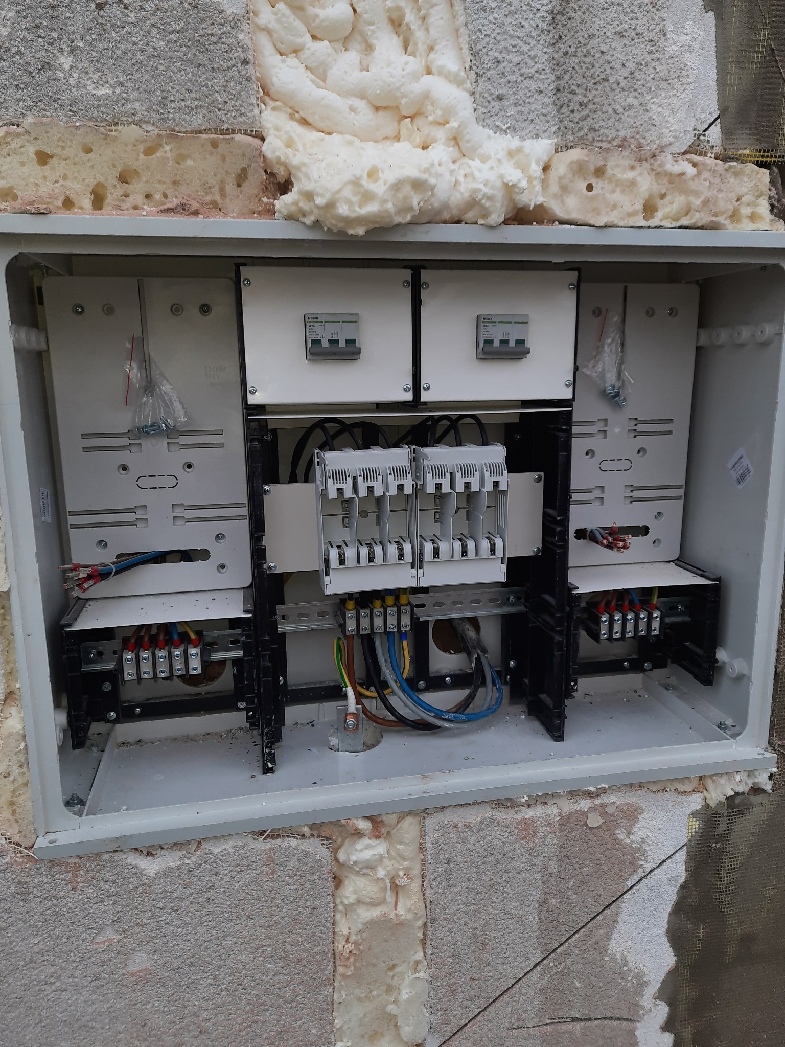 Rozdzielnica elektryczna zewnętrzna, zamontowana na ścianie z widoczną pianką montażową i siatką, z przewodami podłączonymi do bezpieczników i listew zaciskowych.