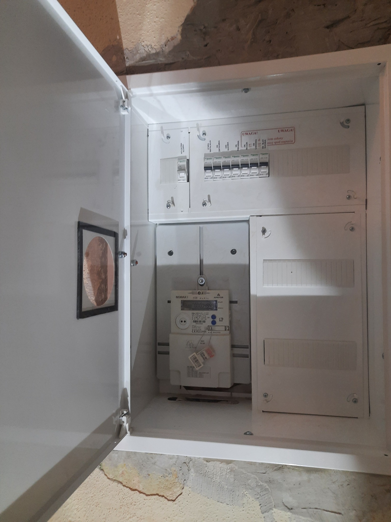 Otwarta skrzynka elektryczna z zamontowanym licznikiem energii elektrycznej i bezpiecznikami, widoczne ostrzeżenia na naklejkach.