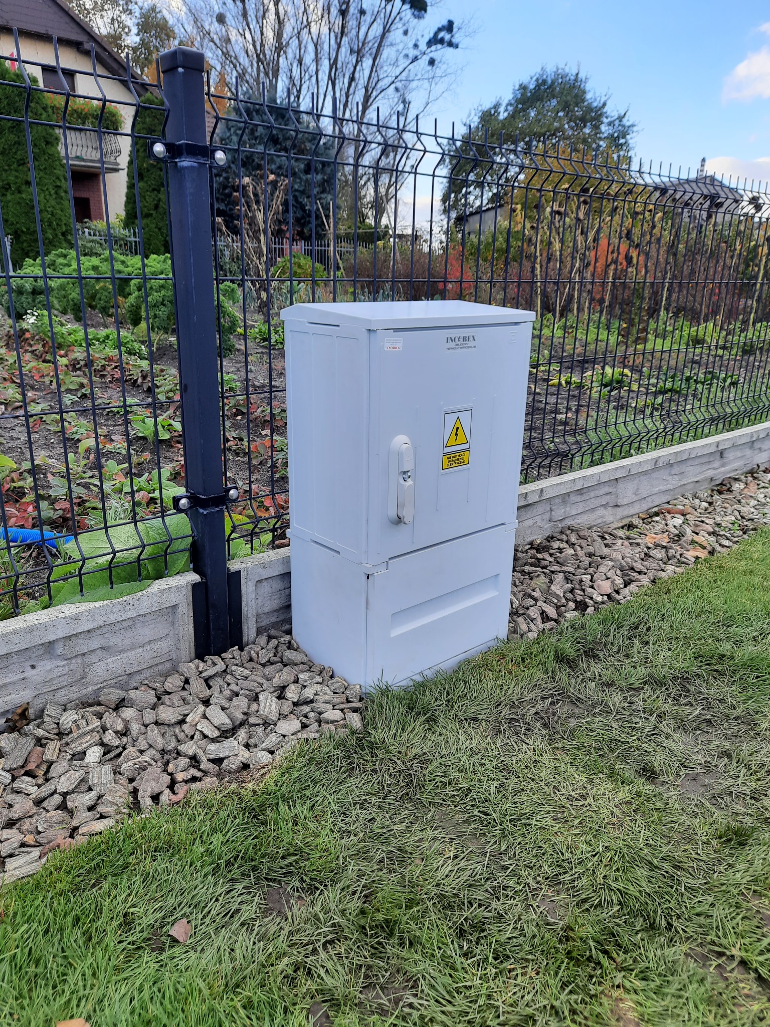 Szara szafa elektryczna IncoBex z ostrzeżeniem o napięciu, umieszczona na trawniku przy ogrodzeniu z siatki.