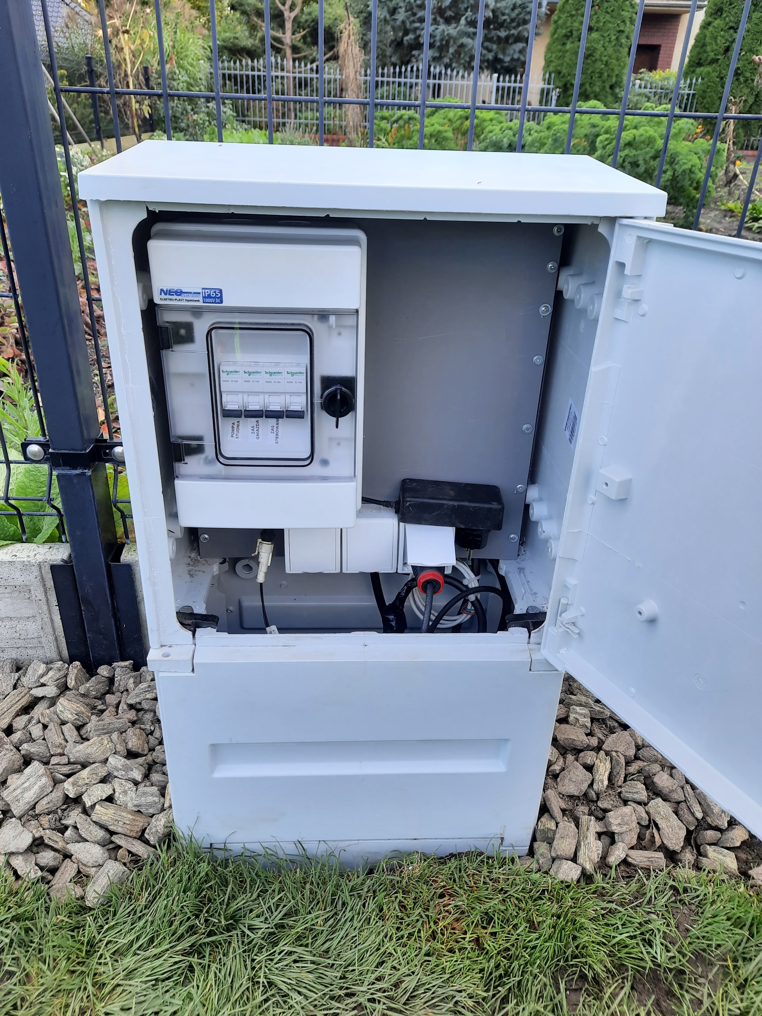 Otwarta, biała szafka elektryczna z zainstalowanymi bezpiecznikami Schneider Electric, widoczna instalacja elektryczna, zamontowana na zewnątrz, przy czarnym metalowym ogrodzeniu, na trawie...