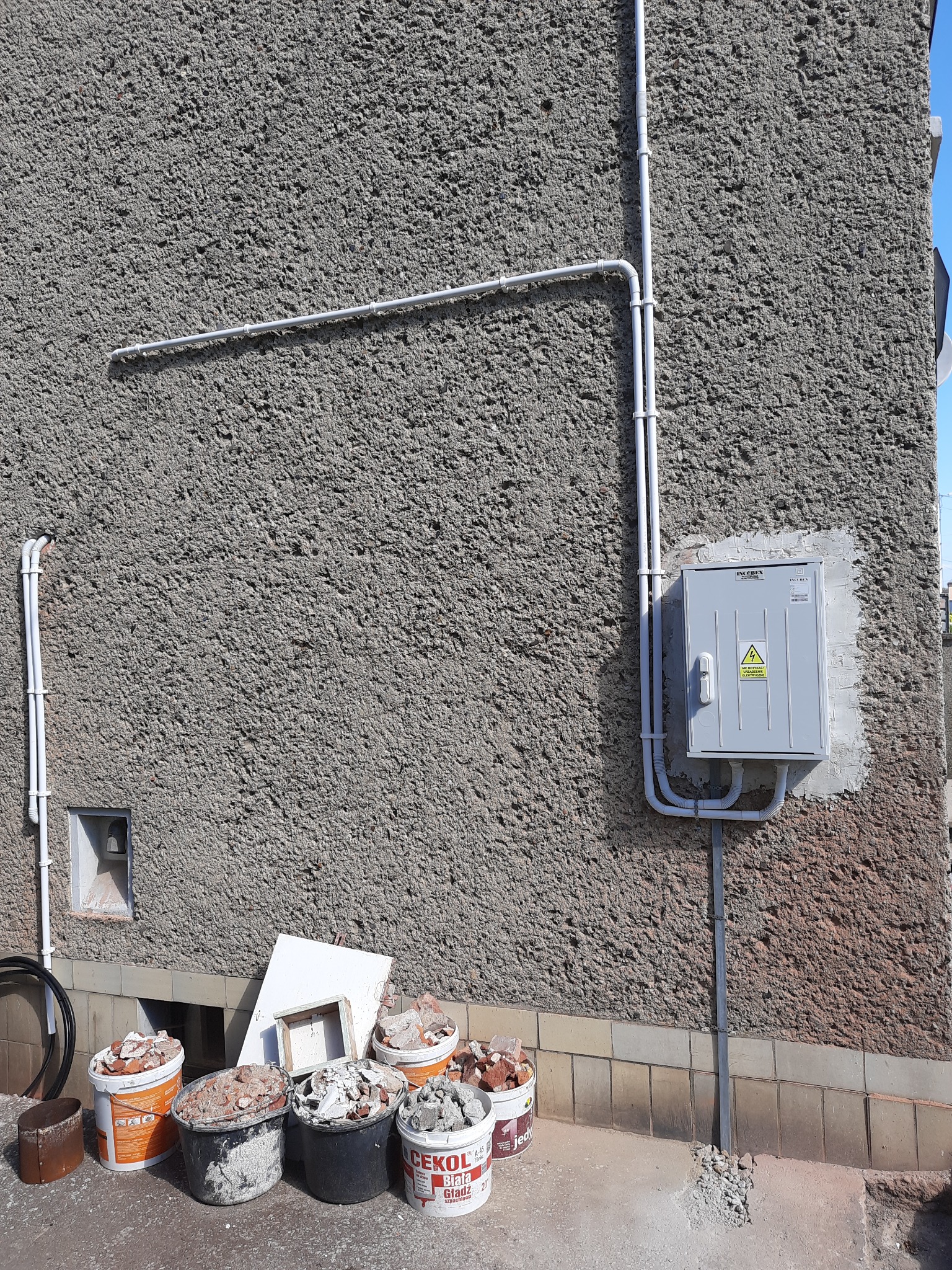 Szara skrzynka elektryczna z symbolem ostrzegawczym wysokiego napięcia zamontowana na chropowatej ścianie budynku, z widocznymi rurami ochronnymi i wiadrami z gruzem u podstawy ściany.