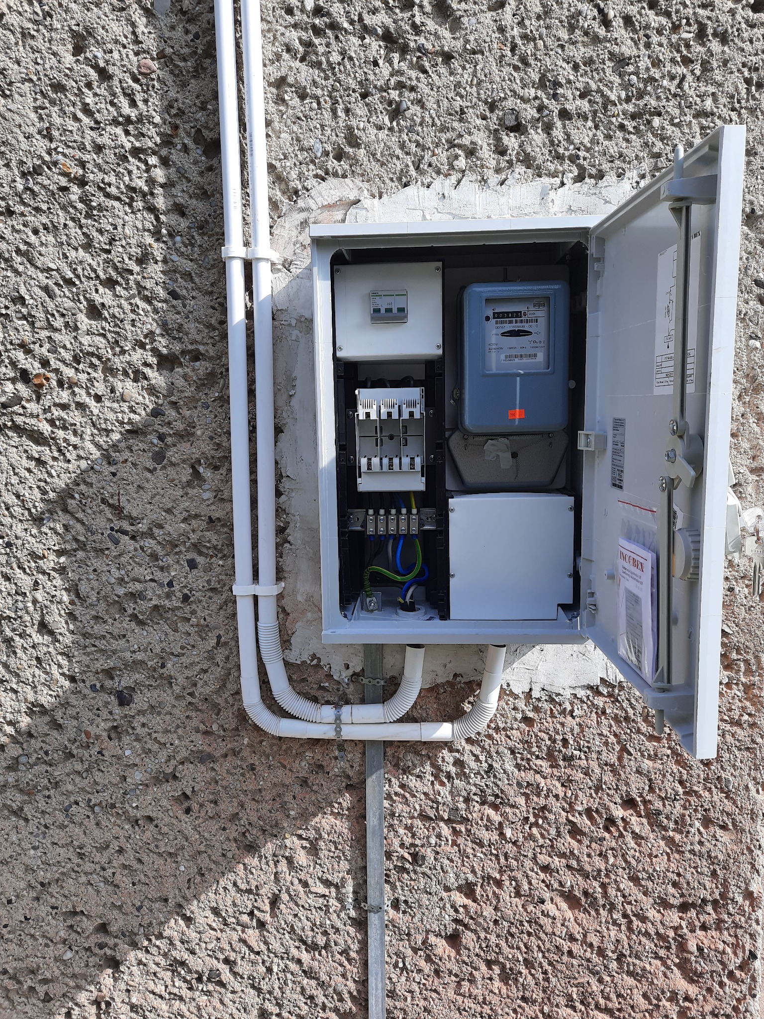 Otwarta skrzynka z licznikiem elektrycznym zamontowana na szorstkiej ścianie zewnętrznej, widoczne przewody, bezpieczniki i instrukcja w opakowaniu foliowym.