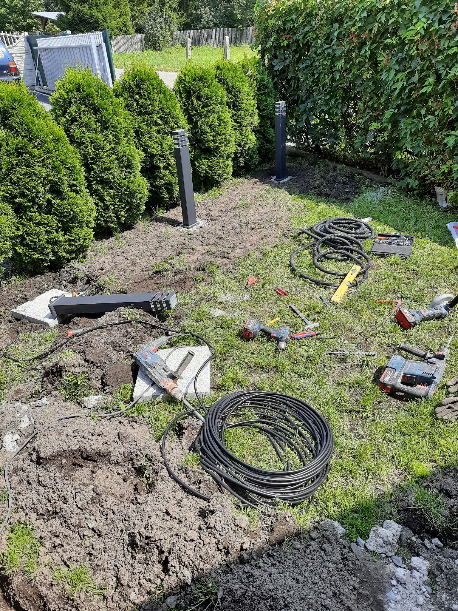 Instalacja oświetlenia ogrodowego: czarne, nowoczesne lampy słupkowe, rozłożone narzędzia (wiertarki, poziomica) i przewody na trawniku podczas montażu.