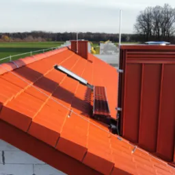 Pomarańczowy dach z dachówki ceramicznej z zamontowaną instalacją odgromową, kominem z blachy w kolorze dachu i oknem dachowym, w tle zielone pole.