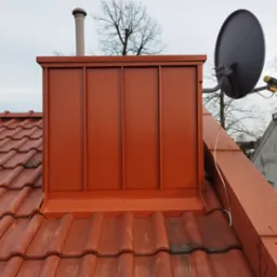 Czerwona, blaszana obudowa komina z anteną satelitarną na tle ceramicznej dachówki i pochmurnego nieba.