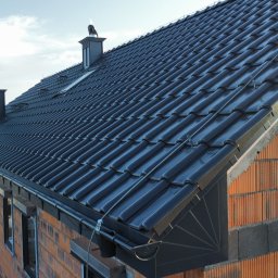 PG - Dachy - Nowy, błyszczący dach z ciemnej dachówki ceramicznej na budynku w trakcie budowy, widoczne kominy i rynny. Ujęcie z boku, w tle krajobraz.