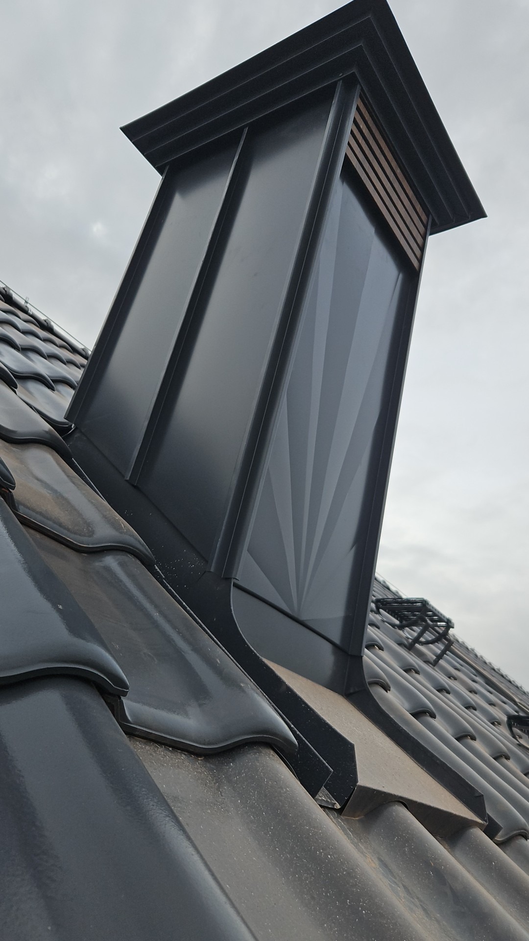 Nowoczesny, grafitowy komin wentylacyjny z blachy na dachu krytym błyszczącą dachówką ceramiczną. Ujęcie z dołu, pochmurne niebo w tle.