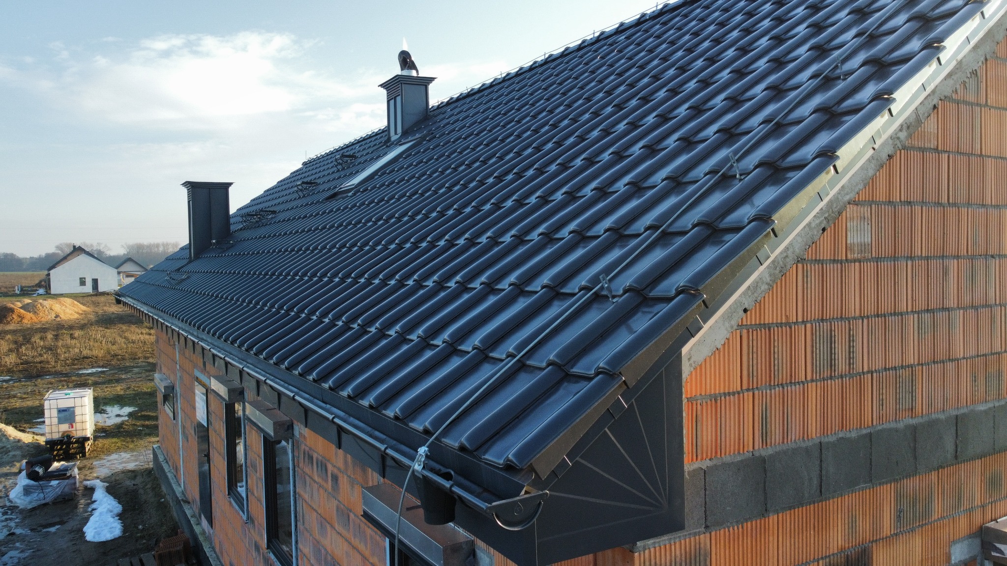 Nowy, błyszczący dach z ciemnej dachówki ceramicznej na budynku w trakcie budowy, widoczne kominy i rynny. Ujęcie z boku, w tle krajobraz.