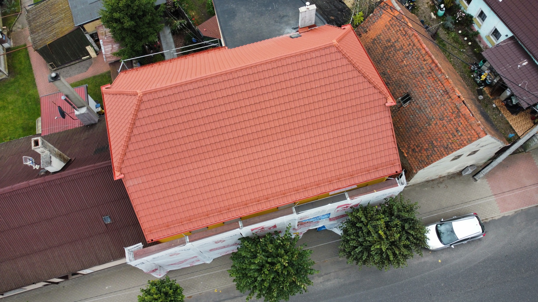Widok z góry na dom z nowym, czerwonym dachem z dachówki ceramicznej. Część elewacji zakryta siatką ochronną. Obok zaparkowany samochód.