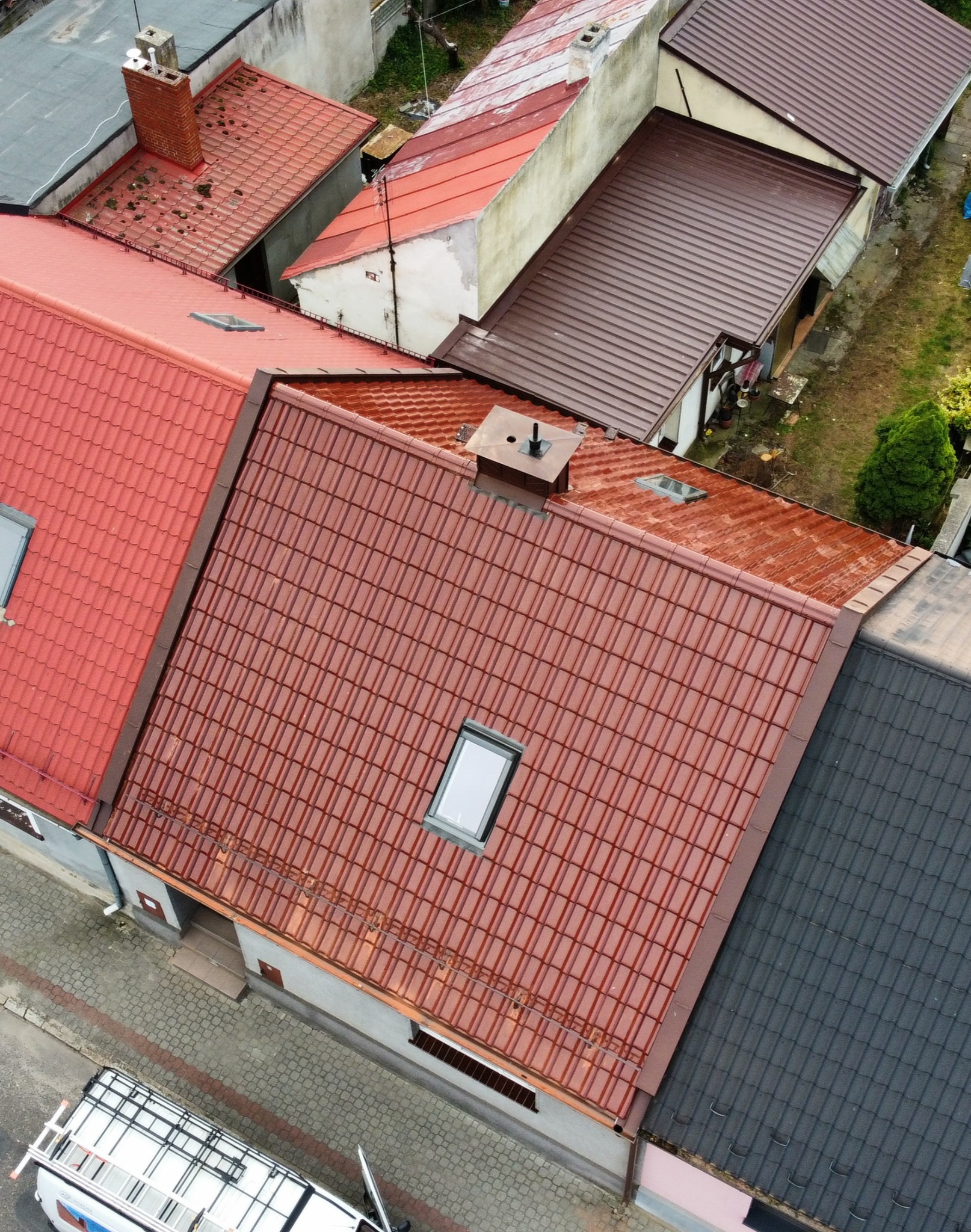 Widok z góry na domy z dachami w odcieniach czerwieni i brązu. Na pierwszym planie dach z oknem dachowym i kominem. W tle widoczny samochód z drabiną.