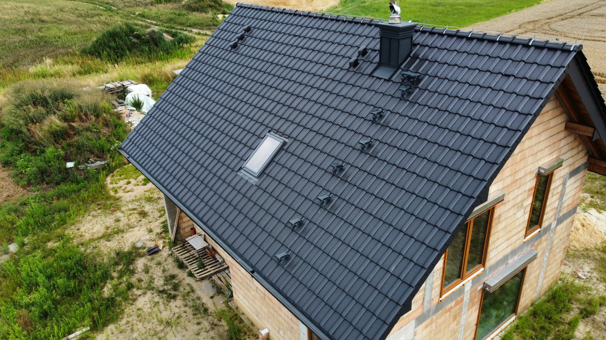 Czarny dach z dachówką ceramiczną, okno dachowe i komin z metalową obróbką, widok z góry na dom w trakcie budowy, otoczony zielenią.