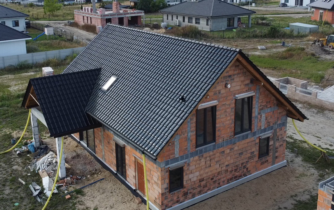 Dom w trakcie budowy z czarnym dachem z dachówki ceramicznej. Widoczny fragment elewacji z cegły i okna z roletami zewnętrznymi. Ujęcie z góry.