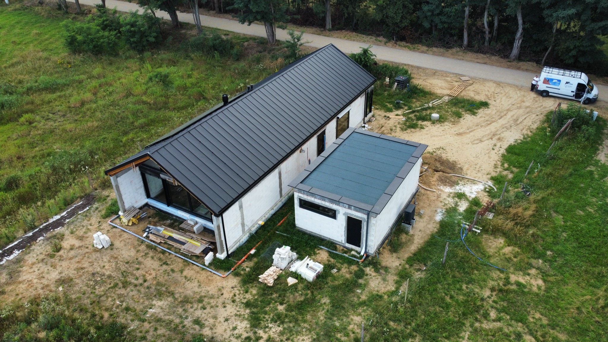 Widok z góry na dom w trakcie budowy z czarnym dachem z blachy trapezowej i płaskim dachem na dobudówce. Na posesji stoi biały bus firmy budowlanej.