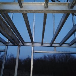 MS STEEL CONSTRUCTION SPÓŁKA Z OGRANICZONĄ ODPOWIEDZIALNOŚCIĄ - Surowa, spawana konstrukcja stalowa z profili, widok pod kątem w górę, na tle ciemnego nieba i drzew bez liści.