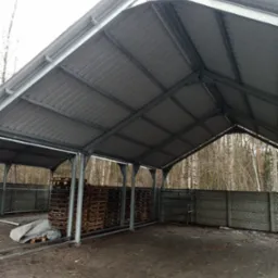 Zadaszona wiata magazynowa o konstrukcji stalowej z blachą falistą, wypełniona paletami drewnianymi, widok z zewnątrz, w tle drzewa i betonowe ogrodzenie.