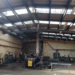 MS STEEL CONSTRUCTION SPÓŁKA Z OGRANICZONĄ ODPOWIEDZIALNOŚCIĄ - Wnętrze warsztatu z dwoma pracownikami przy stanowiskach spawalniczych, widoczne metalowe elementy konstrukcyjne w trakcie obróbki, surowy wystrój ścian i oświetlenie przemysłowe.